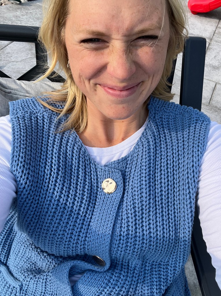 A person smiling while wearing a blue knit sweater with a gold button, sitting outdoors on a patio.