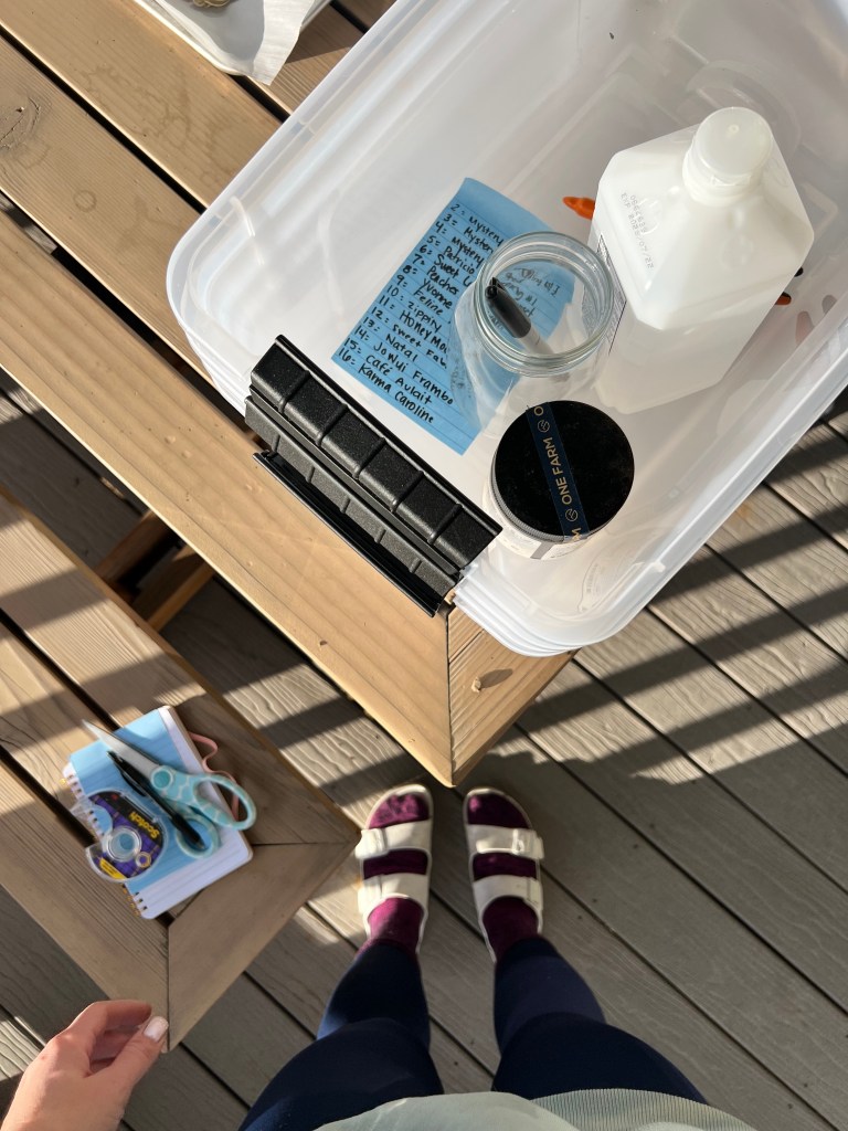A top-down view of a wooden table with a clear plastic container holding various items, including a bottle, a glass jar, and a blue note. To the side, there are a notepad, scissors, and hands reaching towards the container. The person is wearing dark leggings and sandals.