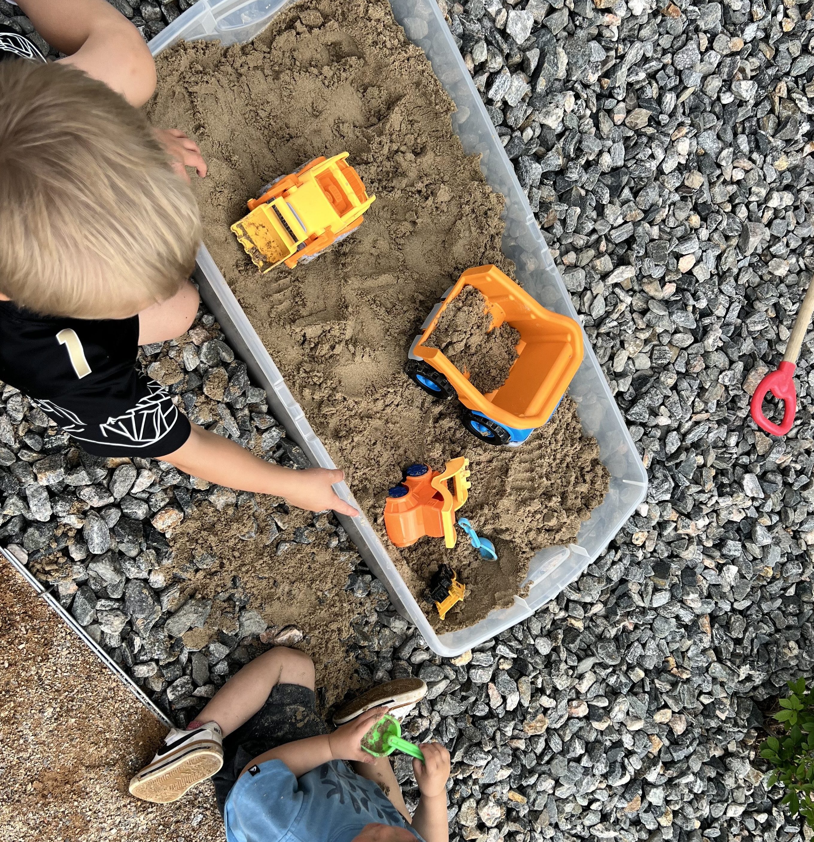 Two young children playing with toy construction trucks in a sandbox, surrounded by gravel.