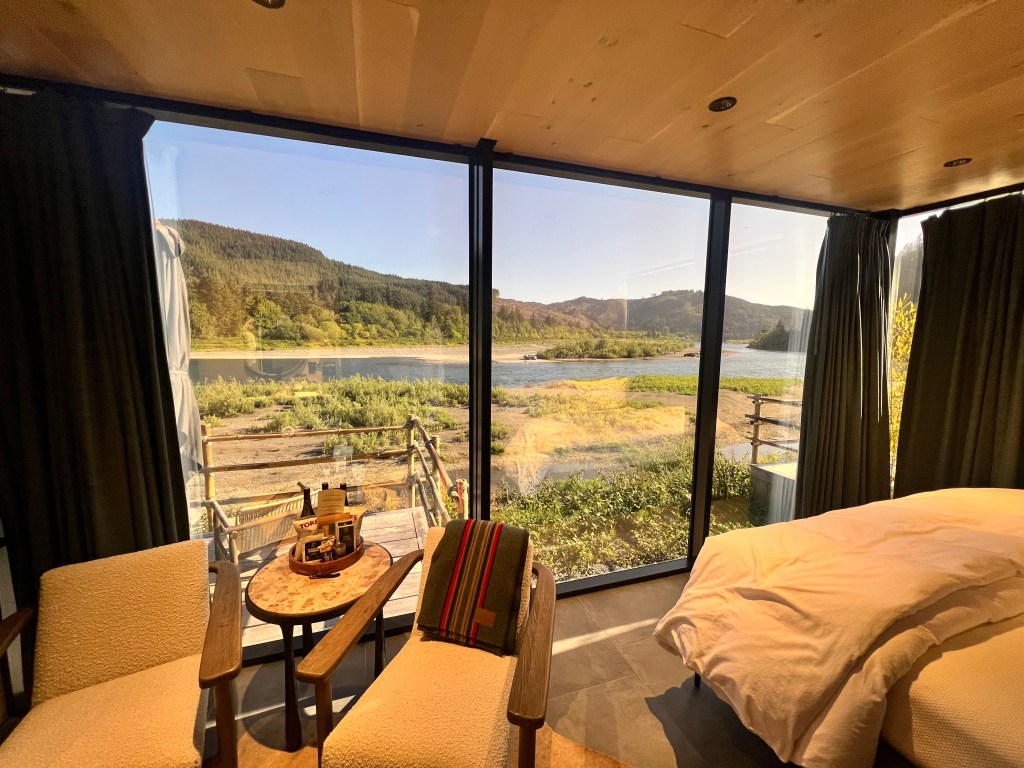 View from a cozy room with two chairs and a table, overlooking a serene river and green landscape surrounded by hills on a sunny day.