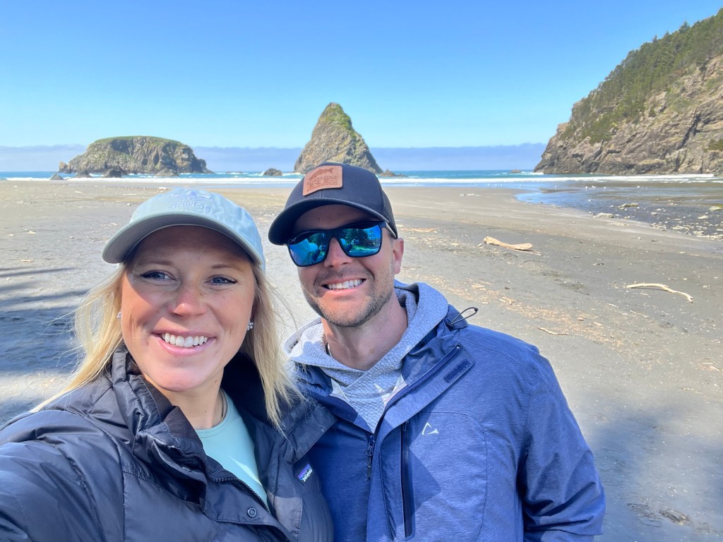 A smiling couple taking a selfie on a beach with rocky outcrops in the background and a clear blue sky.