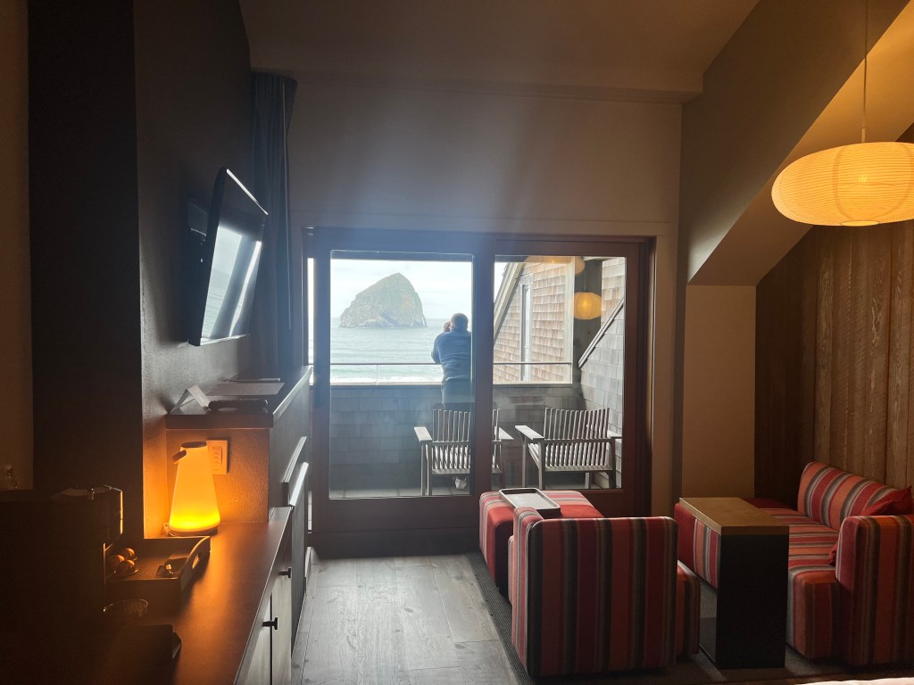 A cozy hotel room with a view of a rocky shoreline, featuring a person standing on the balcony, striped seating area, a TV, and ambient lighting.