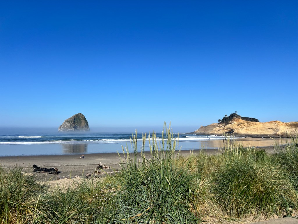 A panoramic view of a beach featuring a prominent rock formation rising from the water, with gentle waves and a clear blue sky.